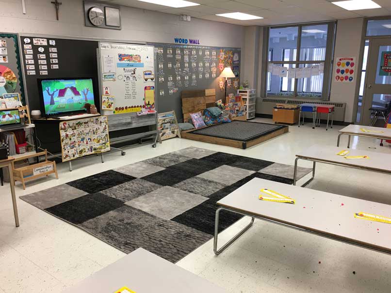 A bright classroom with a reading nook, a checkered rug, and student desks.
