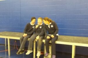 Three students sitting together while the middle student has a microphone as the other two students lean in