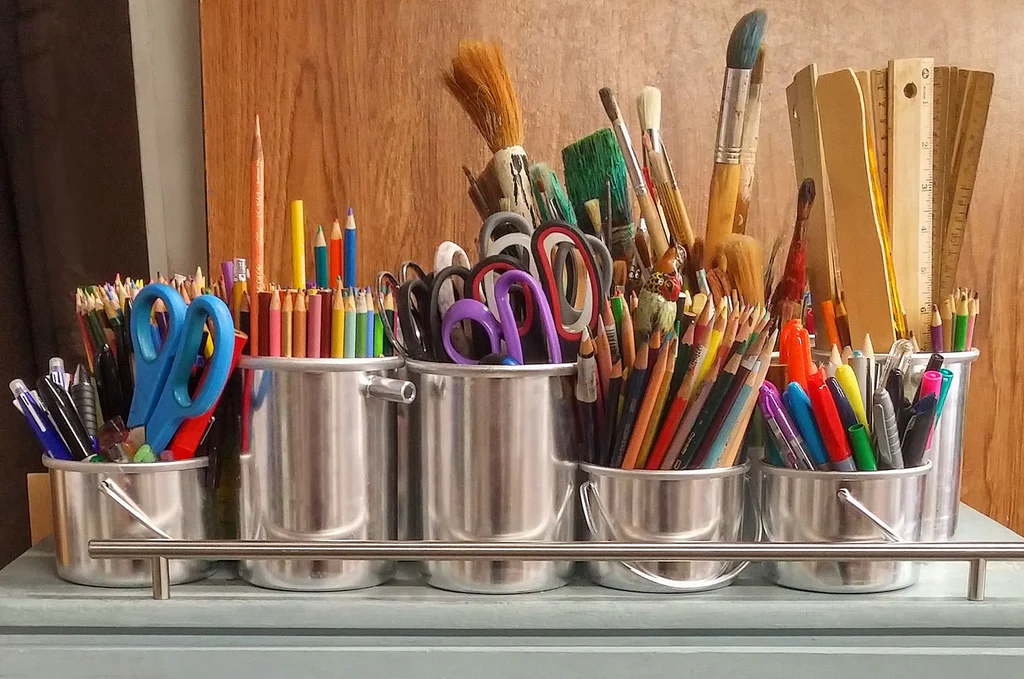 Silver buckets holding art supplies such as rulers, paint brushes, pencil crayons, and scissors