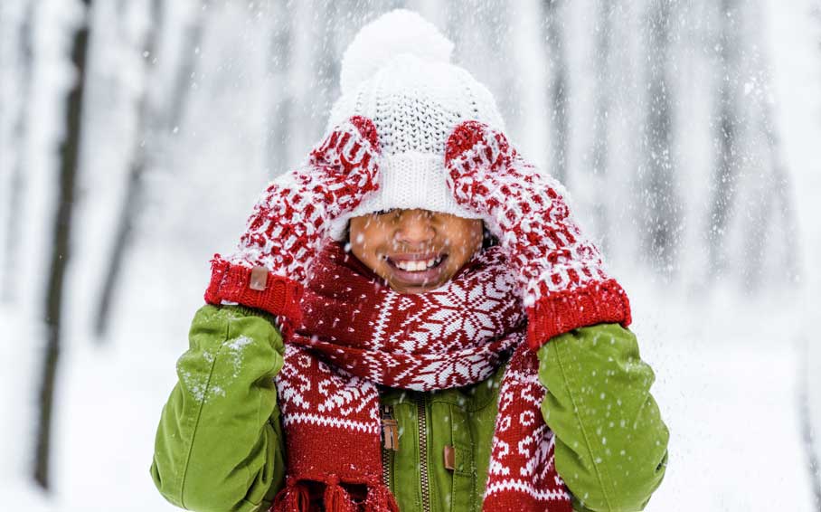 Smiling child outside in the snow wearing a green jacket, white hat and matching red scarf and mittens