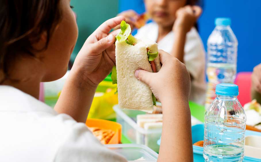 Young girl sitting at a school lunch table with a sandwich in hands